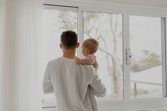 Adult holding a baby while standing by a large window, both looking outside at a softly lit, overcast landscape