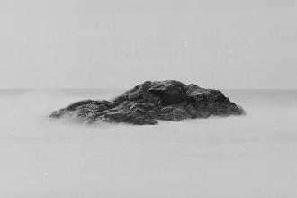 Large dark rock formation emerging from smooth, mist-like water under a pale, overcast sky