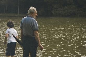 Older adult and child standing at the edge of a calm, reflective body of water, holding items in their hands while looking out across the surface