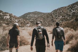Three people walk along a dirt trail surrounded by dry brush, with rocky mountains rising in the background under a clear sky