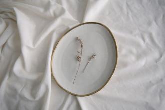 A simple ceramic plate holding two delicate dried sprigs rests on softly wrinkled white fabric