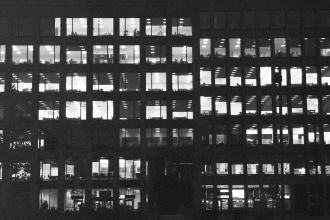 A nighttime view of a large office building, its grid of windows illuminated to reveal people working inside