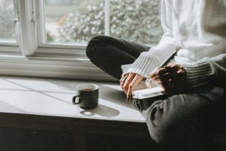 Person sits cross‑legged on a windowsill, writing in a notebook beside a mug of coffee in the sunlight