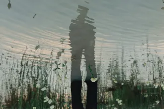 Reflection of a person standing beside rippling water with grass and white flowers, symbolizing Lent’s themes of reflection, prayer and spiritual renewal.