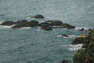 A person wearing a yellow shirt stands on dark rocks surrounded by ocean waves