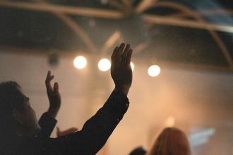 People raise their hands in worship inside a dimly lit room with bright stage lights overhead