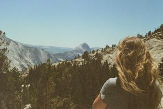 Individual with windblown hair looking out over a mountain landscape filled with pine trees and rugged cliffs