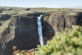A tall waterfall cascades over a steep cliff into a rocky valley surrounded by green terrain