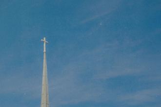 A church steeple with a cross at the top rises into a clear blue sky