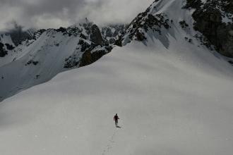 A lone person walks across a wide snowy slope surrounded by towering, rugged mountains and low-hanging clouds