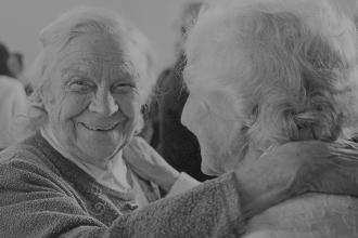 Two older adults embrace closely, one placing a hand gently on the other’s shoulder