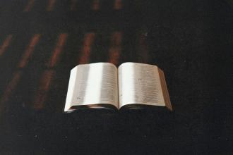 An open Bible rests on a dark surface with warm light softly illuminating the pages