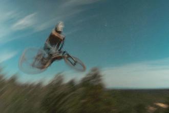 A cyclist performs a high jump on a mountain bike, captured in a motion‑blurred scene against a blue sky
