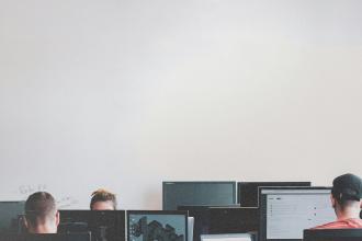 Several people sit at desks working on computers in a bright room with a large blank wall above them