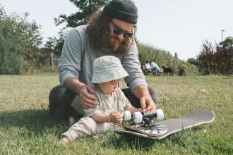 Father helping a baby examine a skateboard while sitting on the grass