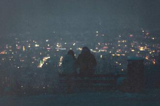 Two people sit on a bench at night overlooking a city filled with glowing lights in the distance