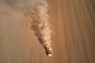 A tractor moves across a large plowed field, leaving a long trail of dust behind it