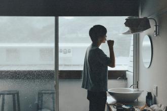 Man brushing their teeth in a small bathroom with a large window overlooking a rainy landscape