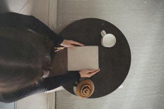 A person sits at a small round table with a closed notebook, a mug of coffee, and a pastry in warm natural light