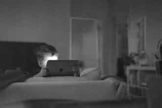 A child sits on a couch in a dim room, holding a tablet with soft light reflecting from the screen