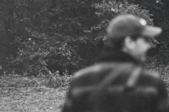 Man wearing a hat, standing in a wooded clearing with dense trees in the background