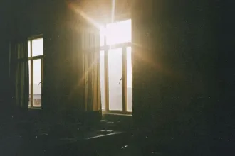 Sunlight streams through tall window panes, casting bright rays and soft golden light across a dimly lit room