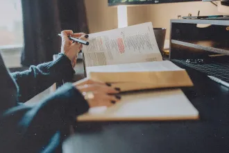 A person studies at a desk with an open book and a Bible, highlighting passages and taking notes in a notebook under soft natural light