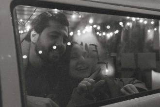 Two people look through a vehicle window adorned with string lights, writing letters in fog