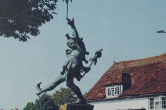 A bronze statue of a lively, dancing figure holding a staff stands in front of a historic building with a red‑tiled roof