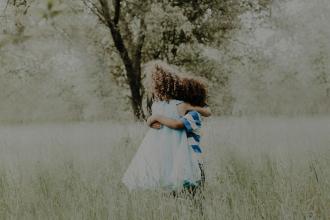 Two children embrace while standing in tall grass beneath trees in a softly lit outdoor setting