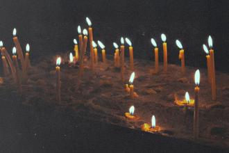 Dozens of thin, glowing candles stand upright in sand, their flames flickering softly in the dark