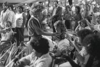 A crowded scene where Gloria Copeland places her hand gently on another person’s head while others around her sit or raise their hands
