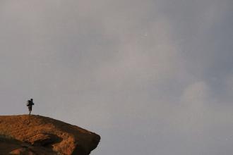 A lone hiker stands on the edge of a rocky cliff under an expansive gray sky