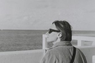 Person standing at a waterfront railing, looking out toward the open sea on a calm, overcast day