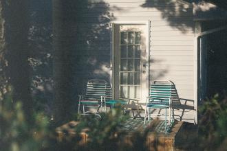 Two striped patio chairs sit on a small wooden porch outside a house, lit by warm late‑afternoon sunlight
