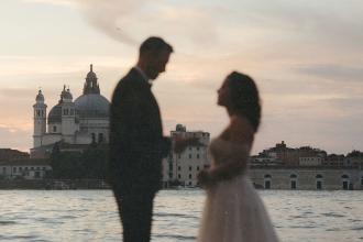 Married couple stand facing each other near the water at dusk, with historic domed buildings in the background and soft pastel colors in the sky