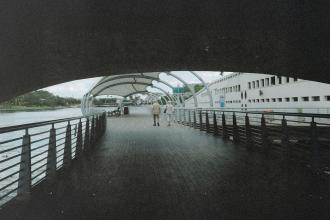 Two people walk together along a covered riverside boardwalk with railings on both sides