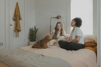 Two people sitting on a bed with two small dogs in a softly lit bedroom