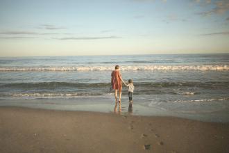 Adult and child standing side by side on the beach, looking out at the calm ocean under a pastel sky