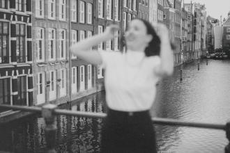 Woman posing on a railing above a waterfront canal, with rows of traditional European buildings in the background.