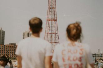 Two people stand outdoors looking toward a tall red metal tower, with buildings and a crowd in the background