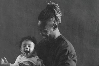 A man smiles while holding a small child in his arms in a softly lit, monochrome portrait