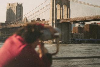 A person looks through a public viewing telescope toward a large suspension bridge crossing over the water