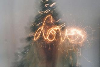 A sparkler writes the word “love” in glowing light trails in front of a blurred Christmas tree