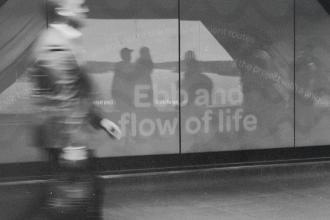 A blurred figure walks past a reflective wall displaying the words “Ebb and flow of life,” with silhouettes visible in the reflection