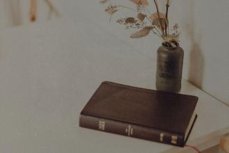 Closed Bible rests on a light-colored table beside a small vase filled with dried branches and leaves
