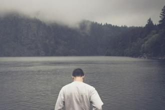 A person stands at the edge of a calm lake surrounded by misty, forested mountains