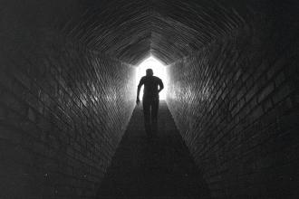 A person walks through a long, dark brick tunnel toward a bright opening at the far end