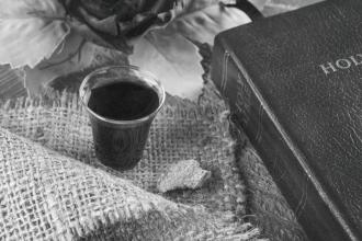 A small cup of communion juice sits on a piece of burlap alongside a broken piece of bread, with a Bible and decorative leaves arranged nearby in a softly lit setting