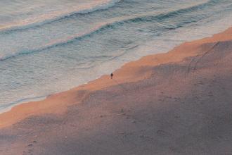 A single person walks along the shoreline at sunrise or sunset, casting a long shadow across the sand as gentle waves roll in